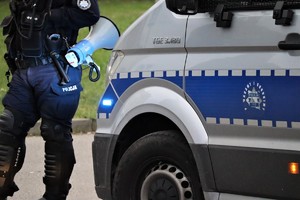 Na zdjęciu umundurowany policjant z megafonem stojący obok radiowozu.