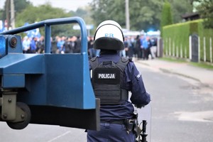 Na zdjęciu umundurowany policjant podczas zabezpieczenia meczu piłki nożnej. W tle grupa kibiców.