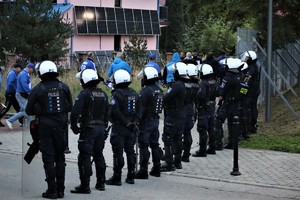 Na zdjęciu umundurowani policjanci podczas zabezpieczenia meczu piłki nożnej. W tle grupa kibiców.