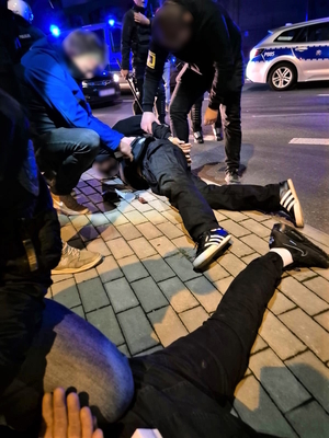 Na zdjęciu mężczyzna leżący na chodniku przytrzymywany przez nieumundurowanych policjantów. Obok nogi drugiego z zatrzymanych mężczyzn. W tle radiowozy policyjne.