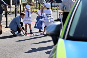 Na zdjęciu dzieci w kaskach z założonymi kamizelkami, które przyklejają znak na drodze.