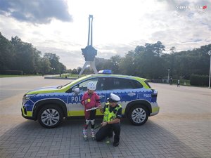 Na zdjęciu radiowóz. Obok radiowozu umundurowany policjant oraz dziewczynka na rolkach trzymająca odblask.