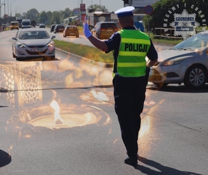 Na zdjęciu umundurowany policjant ruchu drogowego kierujący ruchem na drodze. W tle znicze