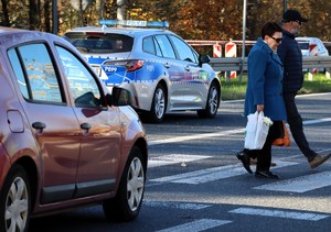 Na zdjęciu radiowóz Policji oraz samochód osobowy w rejonie przejścia dla pieszych, po którym idzie mężczyzna z kobietą.