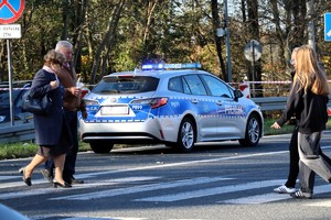 Na zdjęciu radiowóz Policji oraz samochód osobowy w rejonie przejścia dla pieszych, po którym idą piesi.