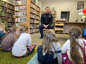Na zdjęciu umundurowany policjant w bibliotece. Przed nim, na dywanie siedzą dzieci