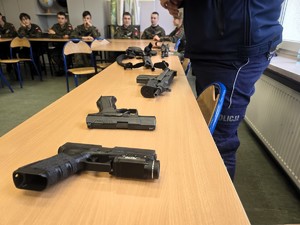 Na zdjęciu różne jednostki broni położone na stole, obok którego stoi umundurowany policjant. W tle uczniowie klasy mundurowej.