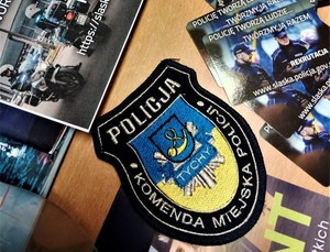 Na zdjęciu policyjne plakaty, ulotki, długopisy i wizytówki zachęcające do pracy w Policji. Pomiędzy nimi naszywka z policyjnego munduru z napisem o treści Komenda Miejska Policji w Tychach.
