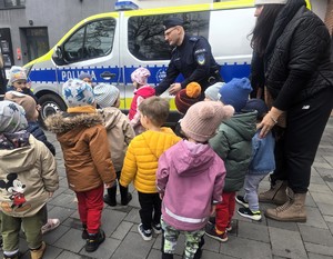 Na zdjęciu umundurowany policjant z policyjną maskotką Sznupkiem oraz grupa dzieci obok radiowozu