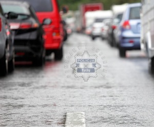 Na zdjęciu jezdnia na której widać samochody stojące w zatorze drogowym. Na środku logo Komendy Miejskiej Policji w Tychach z napisem o treści Pomagamy i chronimy