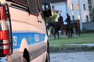 Na zdjęciu oznakowany radiowóz policyjny. W tle policjanci na koniach.