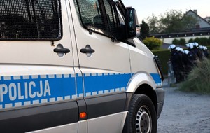 Na zdjęciu policyjny radiowóz. W tle policjanci z oddziałów prewencji.
