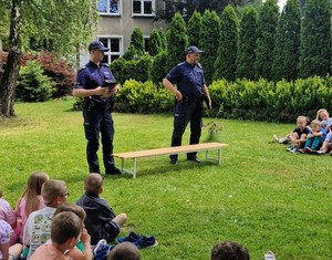 Na zdjęciu policjanci podczas prelekcji. Przed nimi dzieci siedzące na kocach, na trawie