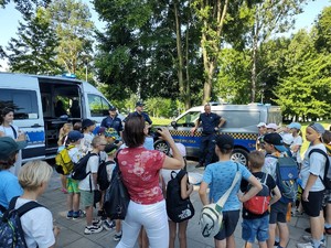 Na zdjęciu grupa dzieci podczas prelekcji policjantów. W tle radiowozy