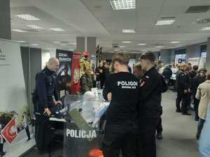 Kolorowe zdjęcie umundurowanego policjanta oraz osób zainteresowanych pracą w Policji. W tle stanowisko promujące formację Policji.