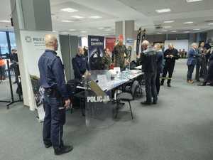 Kolorowe zdjęcie umundurowanego policjanta oraz osób zainteresowanych pracą w Policji. W tle stanowisko promujące formację Policji.