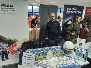 Kolorowe zdjęcie umundurowanego policjanta. W tle stanowisko promujące formację Policji.