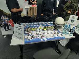 Kolorowe zdjęcie umundurowanego policjanta oraz osób zainteresowanych pracą w Policji. W tle stanowisko promujące formację Policji.