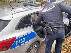 Kolorowe zdjęcie umundurowanych funkcjonariuszy prowadzących sprawcę kradzieży sklepowej do oznakowanego radiowozu.