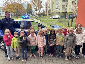 kolorowe zdjęcie dzieci z umundurowanym policjantem przed oznakowanym radiowozem.