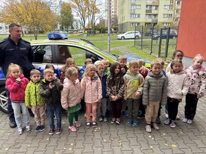 kolorowe zdjęcie dzieci z umundurowanym policjantem przed oznakowanym radiowozem.