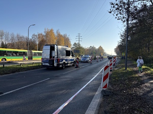 Kolorowe zdjęcie drogi w rejonie cmentarza. Widoczne są zaparkowane oznakowane radiowozy policji, umundurowani policjanci ruchu drogowego kierujący ruchem. Na drodze widoczny odbywający się ruch pieszych oraz pojazdów.