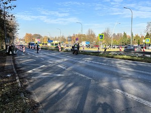 Kolorowe zdjęcie drogi w rejonie cmentarza. Widoczne są zaparkowane oznakowane radiowozy policji, umundurowani policjanci ruchu drogowego kierujący ruchem. Na drodze widoczny odbywający się ruch pieszych oraz pojazdów.