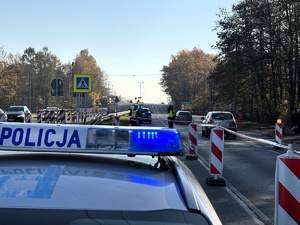 kolorowe zdjęcie policjanta kierującego ruchem na przejściu dla pieszych. W tle widoczny dach radiowozu z włączonymi sygnałami świetlnymi.