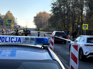 kolorowe zdjęcie policjanta kierującego ruchem na przejściu dla pieszych. W tle widoczny dach radiowozu z włączonymi sygnałami świetlnymi.