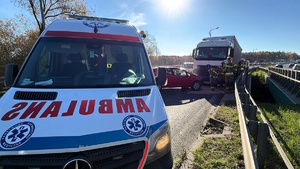 Kolorowe zdjęcie karetki pogotowia. W tle widoczni strażacy przy pojazdach uczestniczących w zdarzeniu drogowym.