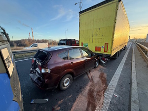 Kolorowe zdjęcie samochodu osobowego, uszkodzonego w wyniku zdarzenia drogowego stojącego na jezdni pomiędzy samochodami ciężarowymi.