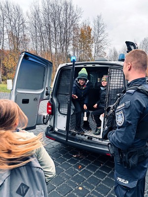 Kolorowe zdjęcie stojącego policjanta oraz dzieci siedzących w oznakowanym radiowozie.
