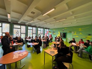 Kolorowe zdjęcie umundurowanego policjanta oraz młodzieży siedzącej przy ławkach