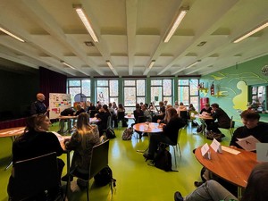 Kolorowe zdjęcie umundurowanego policjanta oraz młodzieży siedzącej przy ławkach