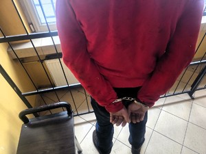 Kolorowe zdjęcie mężczyzny w kajdankach założonych na ręce trzymane z tyłu. W tle kraty pomieszczenia w którym stoi.