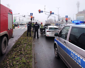 Kolorowe zdjęcie skrzyżowania z sygnalizacją świetlną, na którym widoczne są samochody uczestniczące w zdarzeniu drogowym, oznakowane pojazdy służb oraz umundurowani policjanci.