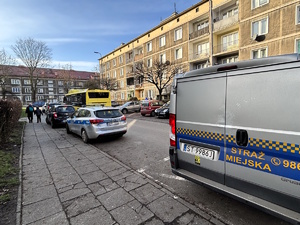 Kolorowe zdjęcie fragmentu drogi w pobliżu budynku wielorodzinnego, na której widoczne są oznakowane radiowozy służb; policji straż miejskiej