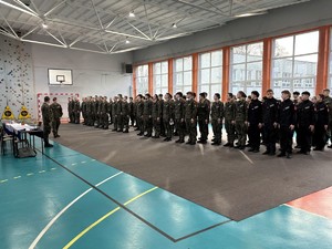 kolorowe zdjęcie umundurowanych drużyn składających się z uczniów klas mundurowych stojących w kolumnach na sali gimnastycznej.