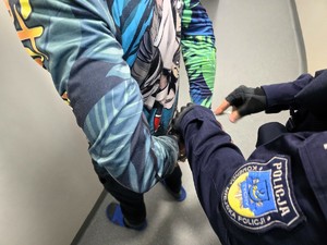 Kolorowe zdjęcie umundurowanego policjanta zakładającego kajdanki na ręce trzymane z tyłu mężczyźnie w kolorowej bluzie.