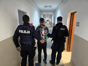 Kolorowe zdjęcie korytarza Komendy Policji, którym umundurowani policjanci prowadzą zatrzymanego mężczyznę.