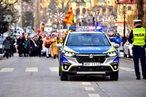 Kolorowe zdjęcie oznakowanego radiowozu Policji oraz umundurowanego policjanta na tle osób biorących udział w orszaku.