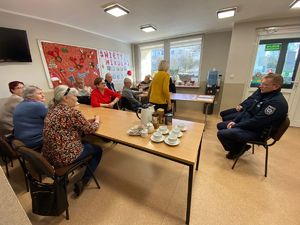 Kolorowe zdjęcie umundurowanego policjanta siedzącego przy stole w otoczeniu siedzących seniorów.