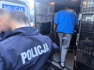 Kolorowe zdjęcie umundurowanych policjantów konwojujących zatrzymanych oznakowanym radiowozem.