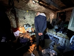 Kolorowe zdjęcie piwnicy w której ukrywali się poszukiwani mężczyźni, w której z stoi zatrzymany z założonymi kajdankami na ręce trzymane z tyłu.