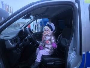 Kolorowe zdjęcie uśmiechniętej dziewczynki która siedzi w oznakowanym radiowozie na miejscu kierowcy w czapce drogówki.