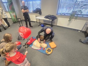 Kolorowe zdjęcie funkcjonariusza Straży Miejskiej szkolącego dzieci z pierwszej pomocy. Na podłodze leży fantom do ćwiczenia pierwszej pomocy.