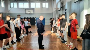 Kolorowe zdjęcie umundurowanego zastępcy Naczelnika Wydziału Prewencji. W tle widoczni uczniowie szkoły.