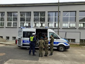 Kolorowe zdjęcie oznakowanego radiowozu policji w otoczeniu uczniów. Obok stoi umundurowany policjant Wydziału Ruchu Drogowego.