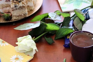 Kolorowe zdjęcie róży leżącej na stole. W tle widać ciasto oraz kubek kawy.