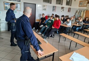 Kolorowe zdjęcie młodzieży uczestniczącej w zajęciach z zasad bezpieczeństwa posługiwania się bronią palną krótką oraz długą. W tle widoczni umundurowani policjanci.